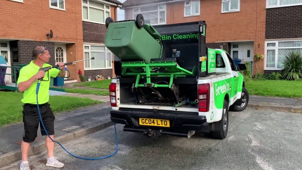 Bin cleaning operative pressure-washing wheelie bins beside a branded green cleaning truck on a residential street